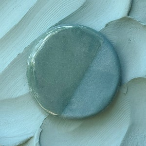 May include: A round, teal-colored ceramic tile with a smooth, glossy finish. The tile has a lighter, almost white, area on one side.