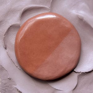 May include: A smooth, round, reddish-brown object with a glossy finish, bisected diagonally by a lighter, pinkish-brown shade. The object rests on a textured, gray surface, possibly clay or a similar material.