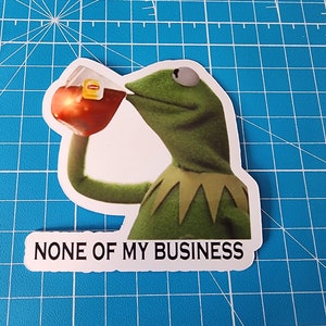 May include: A green frog puppet with a yellow collar is drinking from a glass of iced tea. The text "NONE OF MY BUSINESS" is printed below the image.