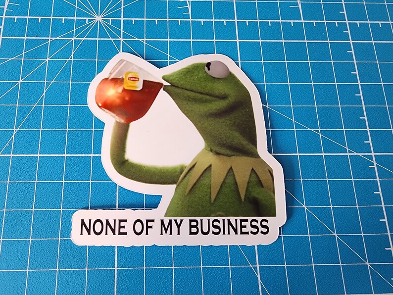 May include: A green frog puppet with a yellow collar is drinking from a glass of iced tea. The text "NONE OF MY BUSINESS" is printed below the image.