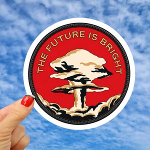 May include: A circular embroidered patch with a red background featuring a beige and black mushroom cloud illustration in the center. The text THE FUTURE IS BRIGHT is stitched in gold thread along the upper curve of the patch border.