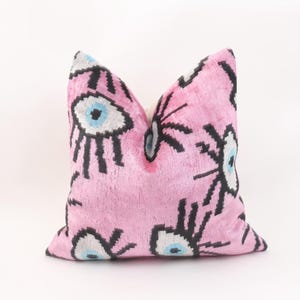 May include: Pink decorative pillow with an eye pattern in black, white, and light blue. The pillow has a soft, velvety texture and is a square shape. The pillow is a home decor item.