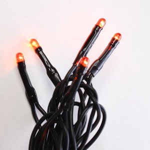 May include: Close-up of a string of orange LED lights on a black wire. The lights are evenly spaced and emit a warm glow. The black wire is tightly wound. The background is plain white.