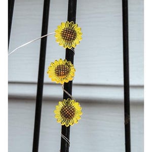 May include: Three yellow sunflower-shaped lights are attached to a black metal rod with silver wire. The sunflowers have a detailed center and are evenly spaced. The background is a white surface with black vertical bars.