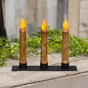 May include: Three brown taper candles with a warm glow on a black metal candle holder.