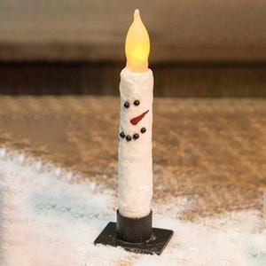 May include: A white taper candle shaped like a snowman with a textured surface, featuring black dot eyes and mouth, an orange carrot-shaped nose, and a yellow flame-shaped LED bulb on top, set in a square black metal base stand.