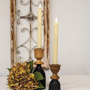 May include: Two cream-colored taper candles in wooden candle holders with black bases. The candle holders have a turned wood design. A bouquet of yellow and green flowers sits in front of a decorative window frame.