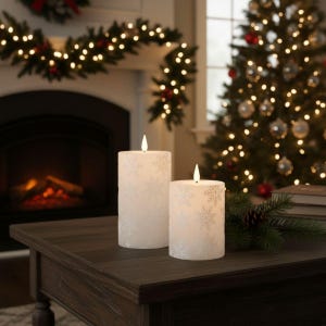 May include: Two white, snowflake-patterned pillar candles with lit flames sit on a dark wooden table. The background includes a fireplace, Christmas tree, and garland, suggesting a festive holiday theme.