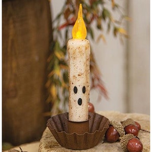 May include: A white candle with a ghost face on it, lit with a yellow flame, sits in a rusty metal candle holder.