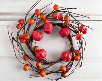 May include: A circular wreath constructed from thin, dark brown intertwined twigs. The wreath is decorated with several small, bright red pumpkins and numerous round, orange and red berries attached to the branches throughout the structure.