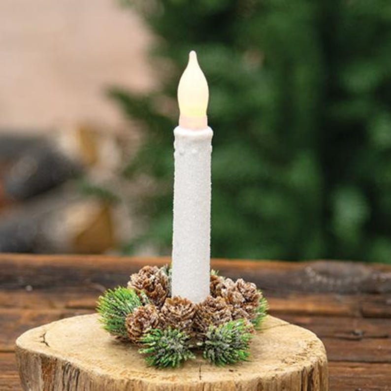 May include: A white, battery-operated candle with a flickering flame effect, surrounded by a wreath of pinecones and greenery. The candle sits on a wooden base, creating a rustic, festive display. Perfect for holiday decor.