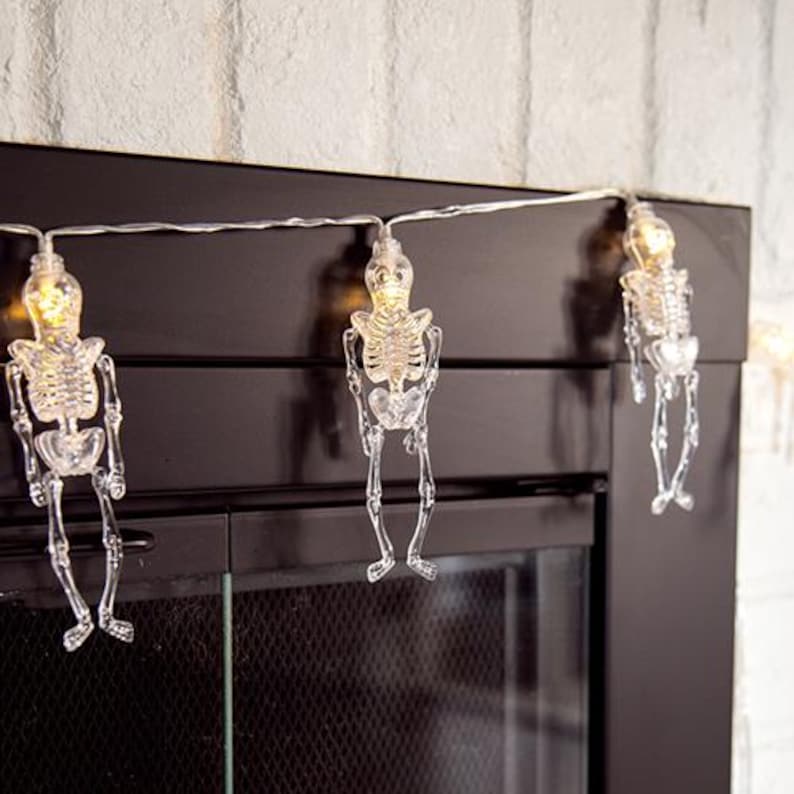 May include: A string of decorative lights featuring three clear, translucent plastic skeleton figures. Each skeleton is illuminated from within by a small, warm-toned light bulb positioned at the top of the skull, connected by a thin, flexible wire.