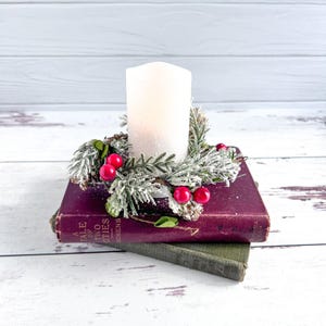 May include: A white pillar candle sits inside a small, frosted wreath made of green pine needles and bright red berries. The candle and wreath are placed on top of a stack of two vintage books, with the top book titled A Tale of Two Cities.