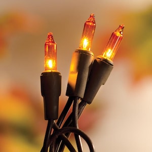 May include: Close-up of three orange-colored Christmas lights with dark green bases and black wires. The lights are lit, emitting a warm glow. The background is a soft blend of orange, yellow, and white.