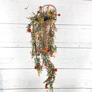 May include: A wall-mounted wicker basket filled with cascading artificial greenery and small, round, burnt-orange flower buds. The basket features a woven texture and a curved handle at the top, with the foliage draping downwards in long stems.