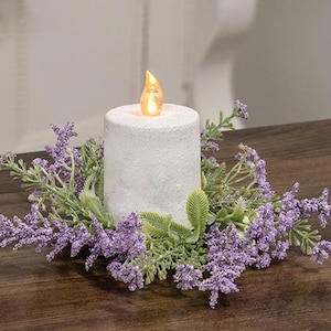 May include: A white candle with a flickering flame sits in a wreath of purple and green flowers. The candle is surrounded by a ring of lavender-colored flowers.