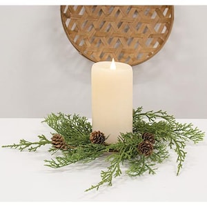 May include: A cream-colored pillar candle with a flickering LED flame, surrounded by a green garland of faux evergreen branches and small pinecones. A woven, circular wall decor is in the background.