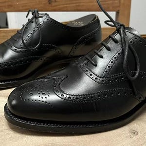 Puede incluir: Un par de zapatos de vestir negros de cuero con cierre de cordones. Los zapatos tienen un diseño de punta de ala y un patrón perforado.