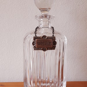 Puede incluir: Una decantador de vidrio transparente con un dise&ntilde;o acanalado y un tap&oacute;n de vidrio. La decantador tiene una etiqueta de metal que dice "whisky" colgando de una cadena.