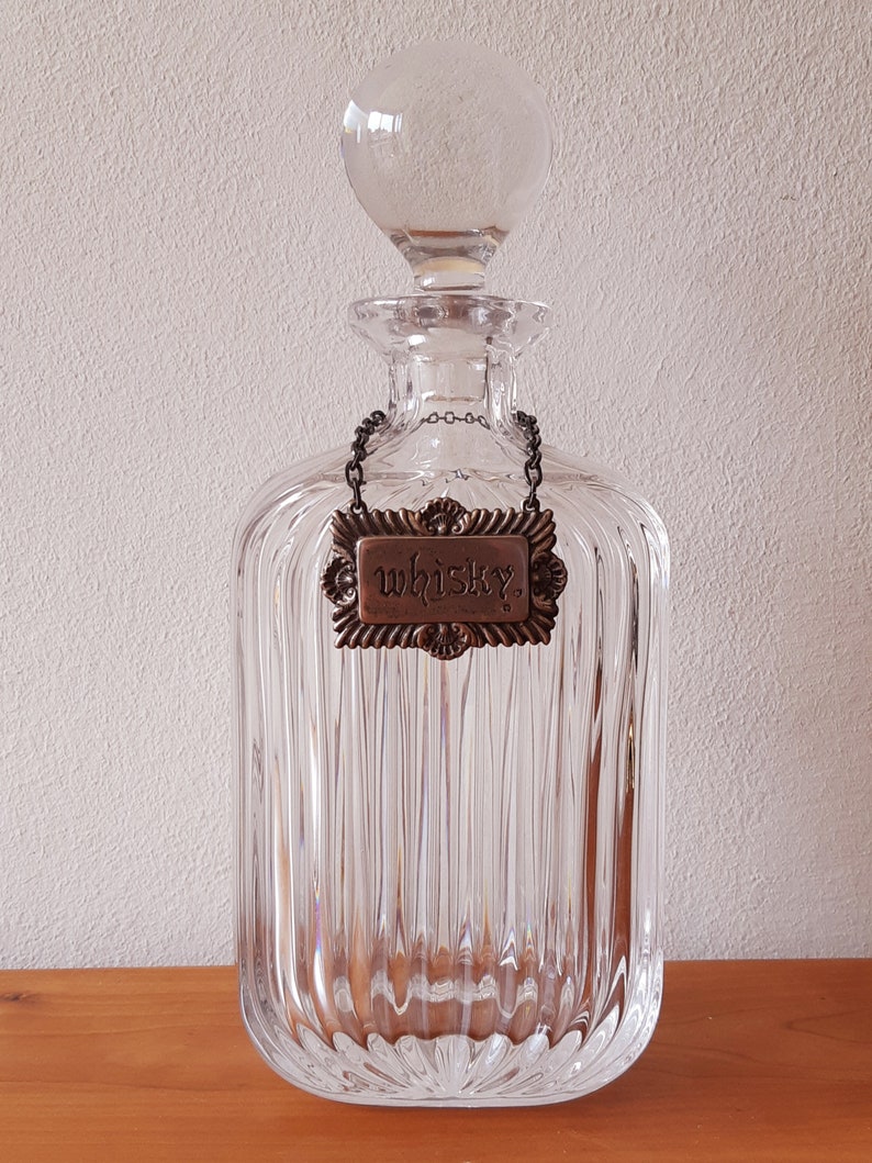 Puede incluir: Una decantador de vidrio transparente con un dise&ntilde;o acanalado y un tap&oacute;n de vidrio. La decantador tiene una etiqueta de metal que dice "whisky" colgando de una cadena.