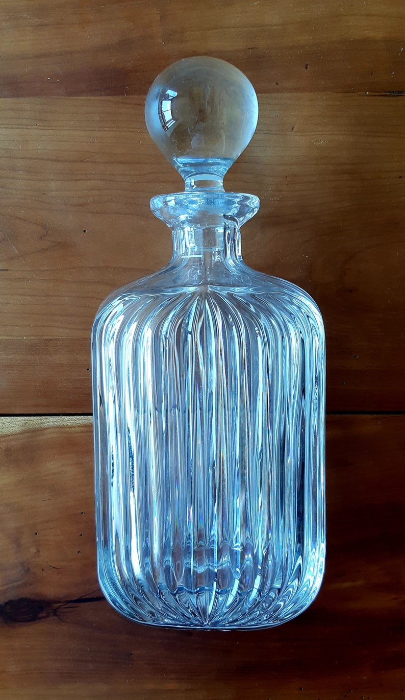 Puede incluir: Una decantador de vidrio transparente con un dise&ntilde;o acanalado y un tap&oacute;n redondo. La decantador est&aacute; vac&iacute;a y tiene un tono azul claro.