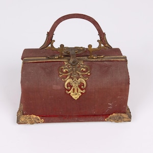 Op de afbeelding: Een vintage stijl toilettas van rood fluweel met gouden metalen accenten. De tas heeft een decoratief handvat en een sluiting.
