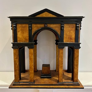 以下が含まれることがあります： 木製の建築模型。クラシックなデザインで、茶色と黒の配色、柱、アーチ、三角形のペディメントが特徴です。小さな正方形の台座が構造体の中心にあります。白い表面に展示されています。