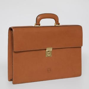 May include: A tan leather briefcase with a top handle and a front flap closure. The briefcase features gold-coloured hardware and a small embossed logo. The leather has a textured finish. The briefcase is approximately 40 cm wide.