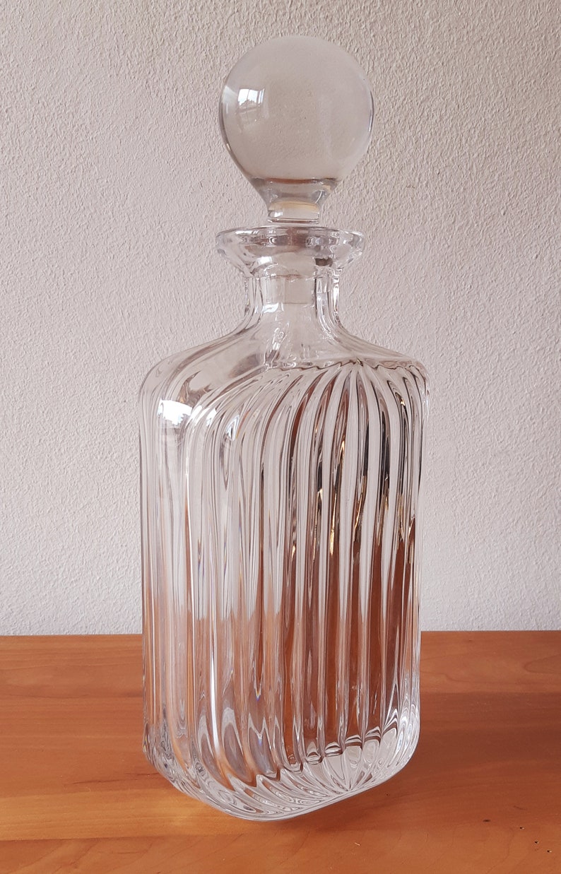 Puede incluir: Una decantador de vidrio transparente con un tap&oacute;n redondo. La decantador tiene un cuerpo cuadrado con nervaduras verticales.