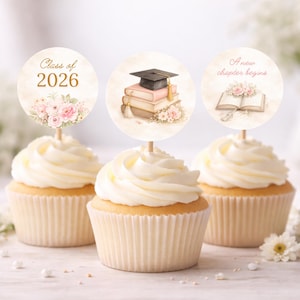 Peut inclure: Trois cupcakes avec glaçage blanc et décorations. Les décorations présentent des motifs sur le thème de la remise de diplômes, notamment le texte "Class of 2026", un chapeau de remise de diplômes sur des livres et la phrase "A new chapter begins". Des motifs floraux sont inclus.