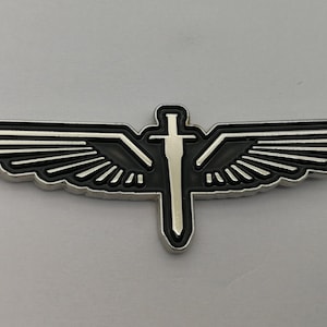 May include: A silver-colored metal pin featuring a sword superimposed over a pair of wings. The wings have a black background with silver-colored lines. The sword is silver-colored with a black outline. The pin is likely a decorative emblem or badge.