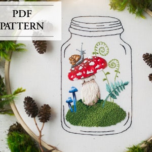 May include: A white embroidery hoop with a hand-stitched design of a glass jar containing a red and white mushroom, a snail, and green ferns. The jar is outlined in black thread. The hoop is surrounded by green moss and pine cones.