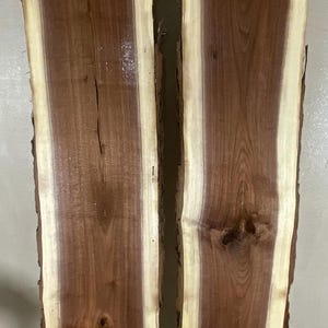 May include: Two adjacent, unfinished wood slabs with a natural edge. The slabs have a rich, dark brown heartwood bordered by a lighter, creamy sapwood. The grain patterns are visible, and the wood appears to be freshly cut.