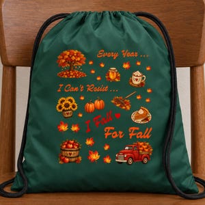 May include: A dark green drawstring bag featuring a colorful autumn-themed graphic with the text Every Year I Can't Resist I Fall For Fall. The design includes a tree, sunflowers, pumpkins, a rake, a slice of pie, a mug, and a red pickup truck.