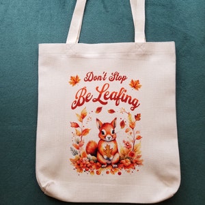 May include: A white tote bag with a squirrel and autumn leaves printed on it. The text "Don't Stop Be Leafing" is printed in red on the bag. The bag is made of 100% polyester linen and is printed on one side.