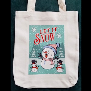 May include: White canvas tote bag with a green, white, and red design featuring a snowman with a pink hat and the text "Let it Snow".
