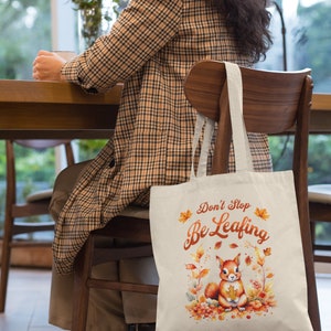 May include: A beige tote bag with a squirrel and autumn leaves design. The bag has the text "Don't Stop Be Leafing" printed on it.