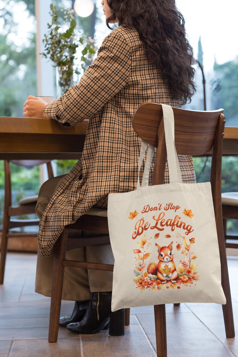 May include: A beige tote bag with a squirrel and autumn leaves design. The bag has the text "Don't Stop Be Leafing" printed on it.