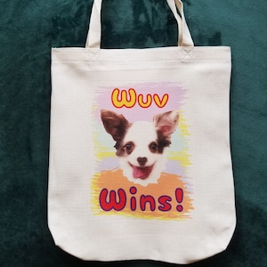 May include: Off-white canvas tote bag with a colorful graphic of a smiling dog. The dog has brown and white fur and a pink tongue. The text "Wuv Wins!" is above and below the dog.