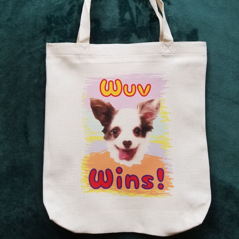 K&ouml;nnte beinhalten: Beige Canvas-Tragetasche mit einer farbenfrohen Grafik eines l&auml;chelnden Hundes. Der Hund hat braunes und wei&szlig;es Fell und eine rosa Zunge. Der Text "Wuv Wins!" steht &uuml;ber und unter dem Hund.
