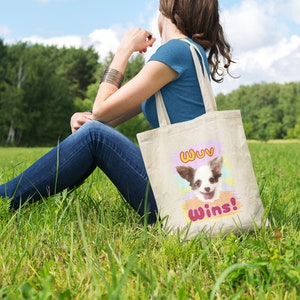 K&ouml;nnte beinhalten: Eine beigefarbene Tragetasche mit einem l&auml;chelnden Chihuahua und dem Text "Wuv Wins!" in einem farbenfrohen Design. Die Tasche wird von einer Person gehalten, die an einem sonnigen Tag in einem grasbewachsenen Feld sitzt.