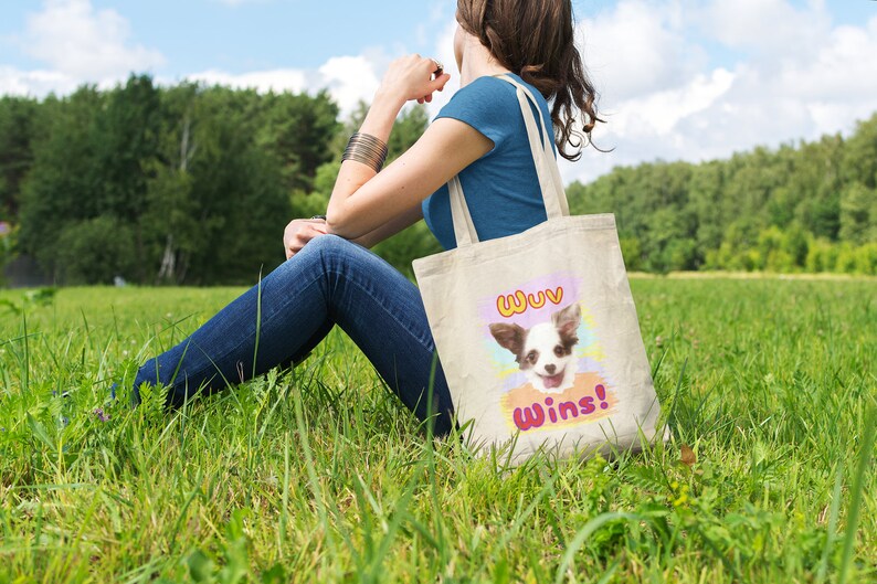 K&ouml;nnte beinhalten: Eine beigefarbene Tragetasche mit einem l&auml;chelnden Chihuahua und dem Text "Wuv Wins!" in einem farbenfrohen Design. Die Tasche wird von einer Person gehalten, die an einem sonnigen Tag in einem grasbewachsenen Feld sitzt.
