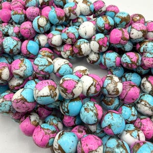 Puede incluir: Primer plano de una colección de cuentas redondas en tonos rosa, azul claro, blanco y dorado. Las cuentas tienen un aspecto moteado y veteado, y están ensartadas. Ideal para la fabricación de joyas.