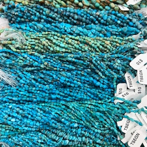 May include: A close-up of a variety of turquoise gemstone beads, ranging in size and shape. The beads are strung on thin cords and are arranged in rows. The image includes price tags with the text "$10 TSEGN" and "TSEGI".