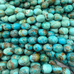 May include: A close-up shot of a pile of turquoise gemstone beads. The beads are oval-shaped and vary in shades of blue and green, with brown and tan veining. The beads are strung together, suggesting they are for jewelry making.