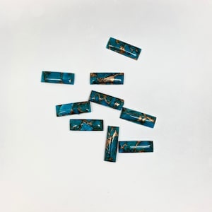 Puede incluir: Una colección de diez cabujones turquesa con motas doradas. Los cabujones son de forma rectangular y tienen una superficie lisa y pulida.