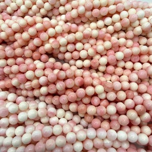 May include: A collection of smooth, spherical beads in varying shades of soft pink, cream, and white. The beads are arranged in multiple strands, creating a dense, textured surface of polished, rounded shapes with subtle color gradients.