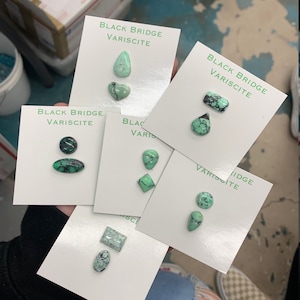 May include: Six cards with the text "Black Bridge Variscite" and various green gemstone cabochons. The cabochons are in different shapes, including teardrop, heart, oval, and square.