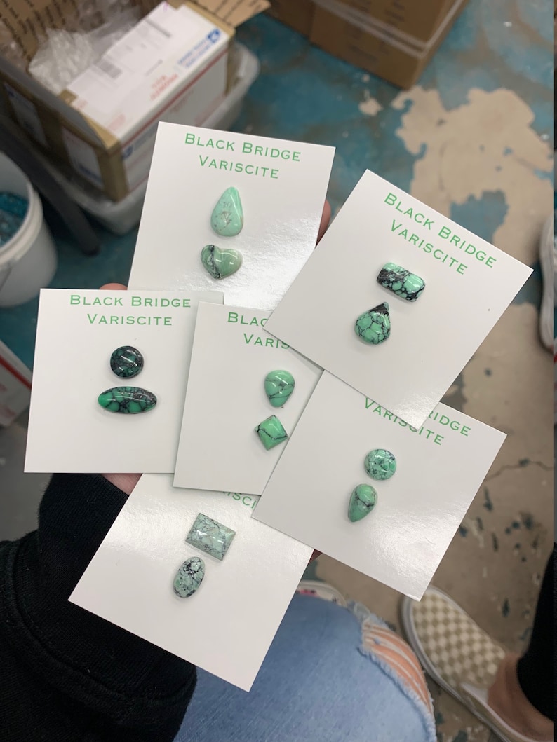 May include: Six cards with the text "Black Bridge Variscite" and various green gemstone cabochons. The cabochons are in different shapes, including teardrop, heart, oval, and square.
