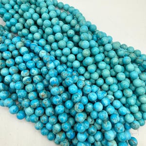 May include: A collection of round turquoise beads arranged in multiple strands. Each bead features a vibrant cyan blue color with natural brown veining patterns throughout the surface. The beads are smooth, polished, and strung together on thin white threads.